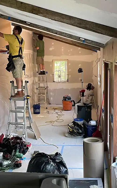 Kitchen under renovation with exposed drywall, construction tools, and workers on ladders.